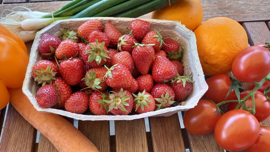 Erdbeeren in einer Pappschale stehen auf einem Holztisch. Daneben liegt anderes Obst und Gemüse.