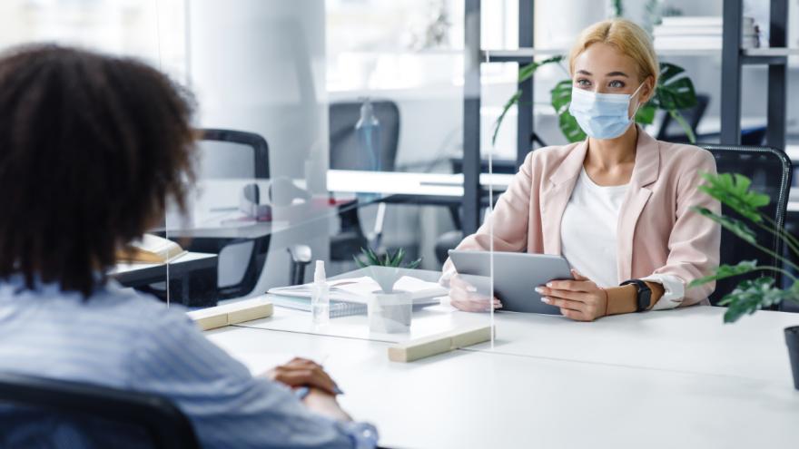 Zwei Frauen sitzen sich bei einer Beratung gegenüber, tragen Schutzmasken und haben eine transparente Scheibe zwischen sich.