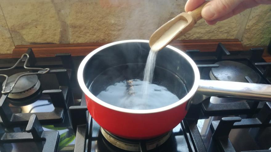Ein roter Topf mit heißem Wasser steht auf einem Gasherd. Eine Hand gibt mit einem Holzlöffel Salz in das Kochwasser.