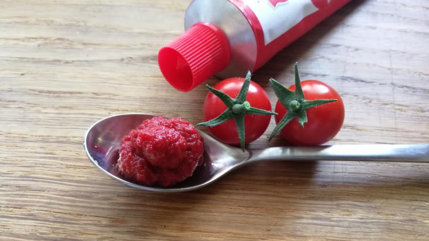 In einem Löffel befindet sich Tomatenmark; daneben eine Tube mit Tomatenmark und zwei kleine Cocktailtomaten.