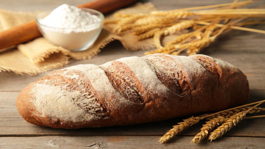 Ein länglicher Laib Brot liegt auf einem Holztisch, daneben liegen Ähren und einen kleine Schüssel Mehl steht auf dem Tisch.. 