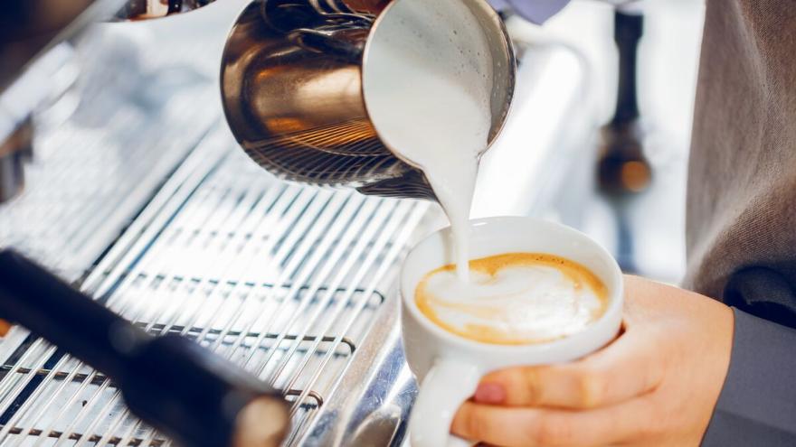 Eine Person gießt aus einer Edelstahlkanne aufgeschäumte Milch in eine Cappuchinotasse