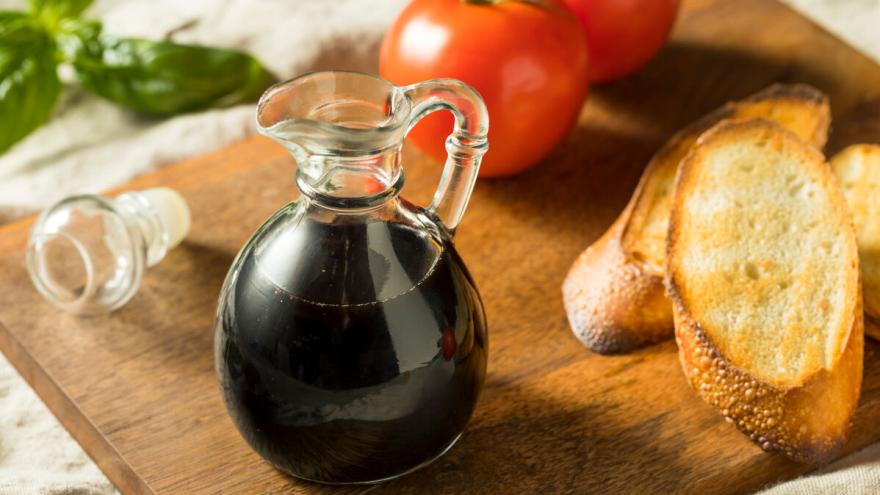 Ein Glaskännchen mit Balsamico Essig steht auf einem Holzbrett, daneben liegen zwei Scheiben getoastetes Baguette und zwei Tomaten