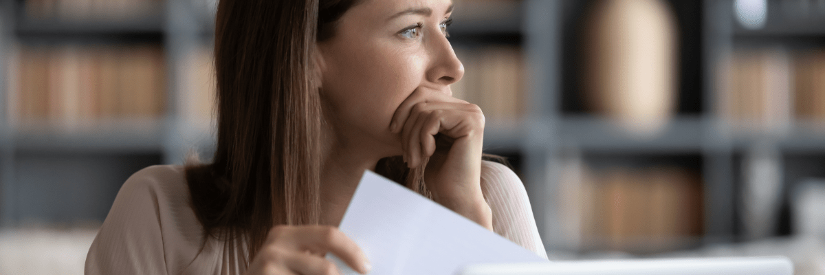 Eine junge Frau sitzt am Schreibtisch, hält einen Brief in der Hand und blickt nachdenklich.